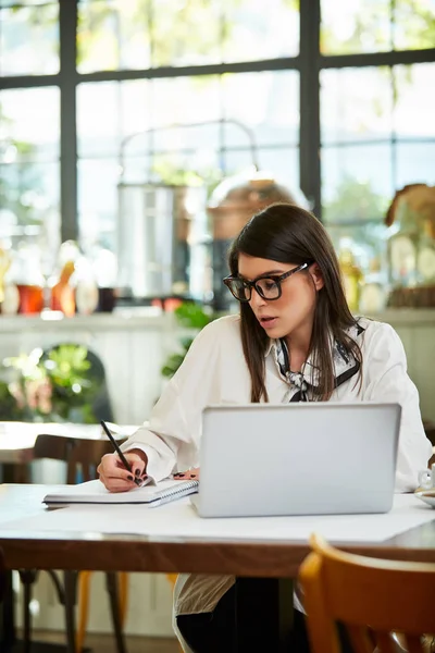 Joven empresaria atractiva positiva en los años treinta vestida con camisa sentada en la cafetería y tareas de escritura en la agenda. En la mesa está el portátil. Concepto de negocio remoto . —  Fotos de Stock