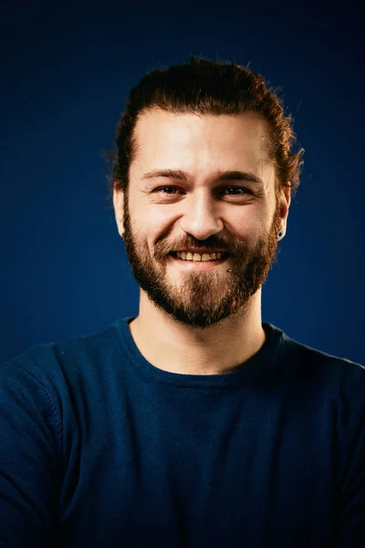 Young handsome smiling bearded caucasian hipster in blue sweater standing in front of blue background and looking at camera. — Stock Photo, Image