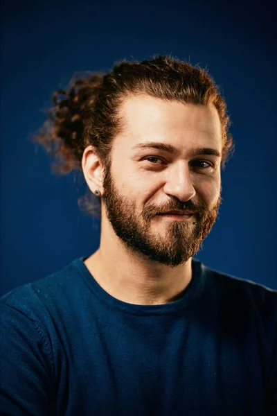 Young charming attractive Caucasian bearded hipster with curly hair dressed in sweater standing in front of blue background and looking at camera. — 스톡 사진