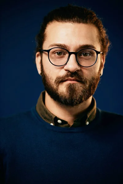 Young attractive caucasian serious bearded hipster with eyeglasses standing in front of blue background. — Stock Photo, Image