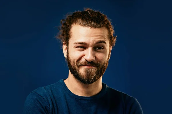 Young attractive caucasian bearded hipster with curly hair dressed in sweater standing in front of blue background with one eyebrow raised and looking away. — Stock Photo, Image