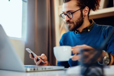 Genç, sakallı hippi evinde oturmuş elinde bir fincan kahveyle akıllı telefon kullanıyor..