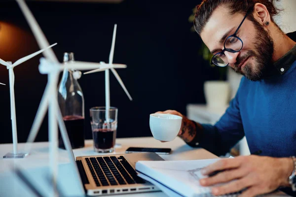 Jonge innovatieve, toegewijde bebaarde medewerker zit in zijn moderne kantoor en tekent schetsen van windmolens en drinkt koffie. Duurzame ontwikkeling. — Stockfoto