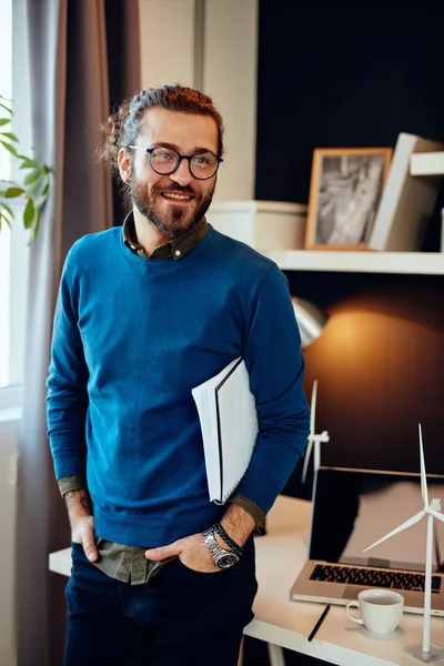 Guapo positivo joven hipster caucásico de pie en su oficina, sosteniendo el papeleo bajo la axila y preparándose para ir a reunirse con clientes importantes . — Foto de Stock