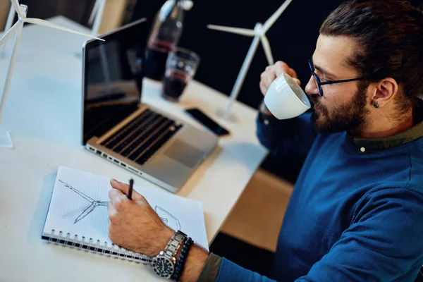 Jonge innovatieve, toegewijde bebaarde medewerker zit in zijn moderne kantoor en tekent schetsen van windmolens en drinkt koffie. Duurzame ontwikkeling. werknemer zit in zijn moder — Stockfoto
