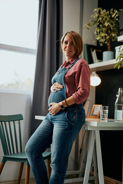 Stolt vacker kaukasisk framtida mor lutar sig på skrivbordet i sitt hemmakontor och rör vid magen medan du tittar på kameran. — Stockfoto