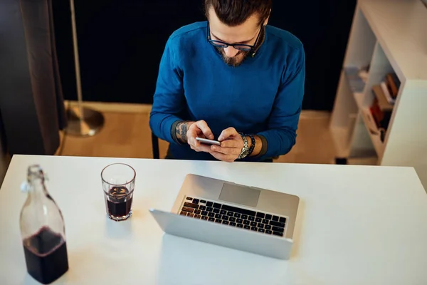 Ελκυστικός καυκάσιος γενειοφόρος επιχειρηματίας που κάθεται στο γραφείο του, χρησιμοποιεί έξυπνο τηλέφωνο και κάνει ένα διάλειμμα από την εργασία. — Φωτογραφία Αρχείου