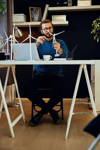 Joven ingeniero barbudo caucásico innovador sentado en su oficina moderna y sosteniendo el modelo de molino de viento. Concepto sostenible . — Foto de Stock