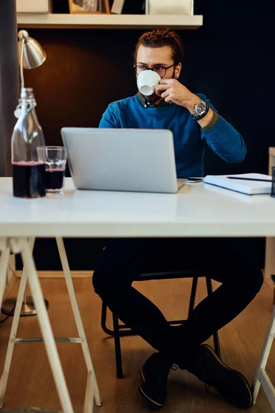 Νεαρός ελκυστικός καυκάσιος γενειοφόρος ελεύθερος επαγγελματίας κάθεται στο γραφείο του και πίνει πρωινό καφέ. Στο τραπέζι είναι laptop. — Φωτογραφία Αρχείου