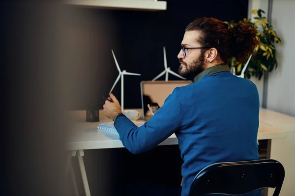 Joven e innovador empleado barbudo dedicado sentado en su moderna oficina y dibujando bocetos de molinos de viento. Concepto de desarrollo sostenible. — Foto de Stock