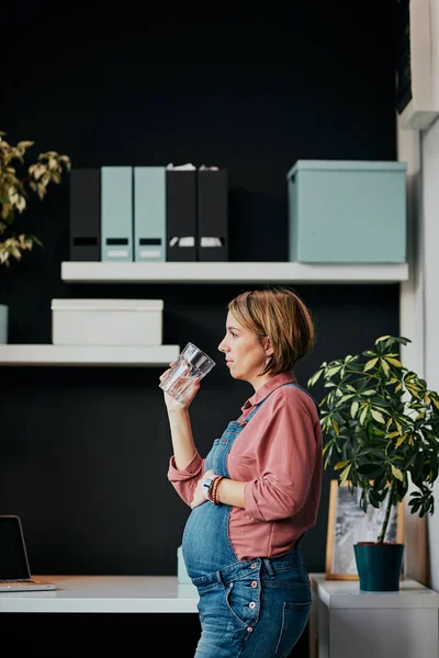 Bedårande kaukasisk gravid frilansare som står på sitt kontor, rör vid magen, dricker vatten och tar en paus från jobbet. — Stockfoto