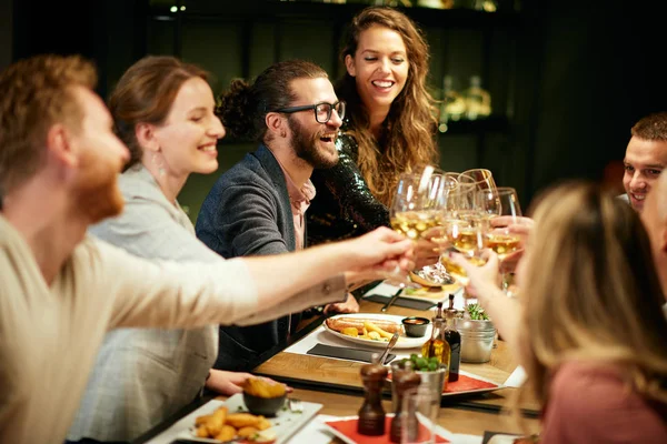 Beste Freunde sitzen zum Abendessen im Restaurant und stoßen mit Weißwein an. Auf dem Tisch liegt Essen. Stockbild