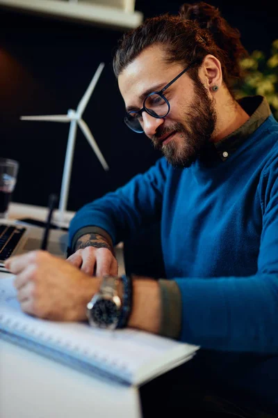 Jonge innovatieve bebaarde medewerker zit in zijn moderne kantoor en tekent schetsen van windmolens. Duurzame ontwikkeling. — Stockfoto