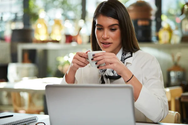 Atractiva morena elegante caucásica sentada en la cafetería, bebiendo café y mirando el portátil. En la mesa está el portátil . — Foto de Stock