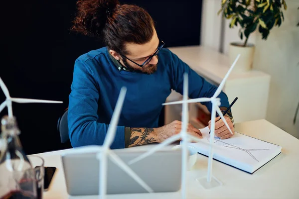 Молодой инновационный преданный бородатый работник сидит в своем современном офисе и рисует наброски ветряных мельниц. Концепция устойчивого развития. — стоковое фото