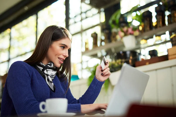 Atractiva morena caucásica sonriente en suéter sentado en la cafetería, tecleando en el teclado y sosteniendo la tarjeta de crédito . — Foto de Stock
