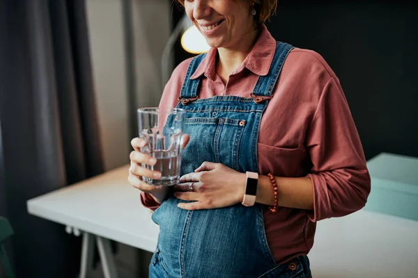 Bedårande kaukasisk gravid frilansare som står på sitt kontor, rör vid magen, dricker vatten och tar en paus från jobbet. — Stockfoto