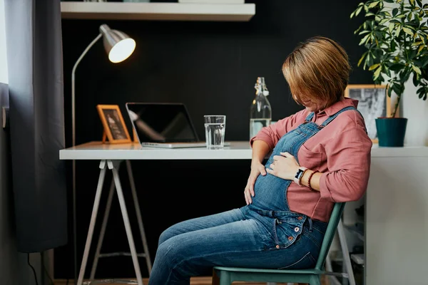Proud future mother sitting in her home office and touching belly.