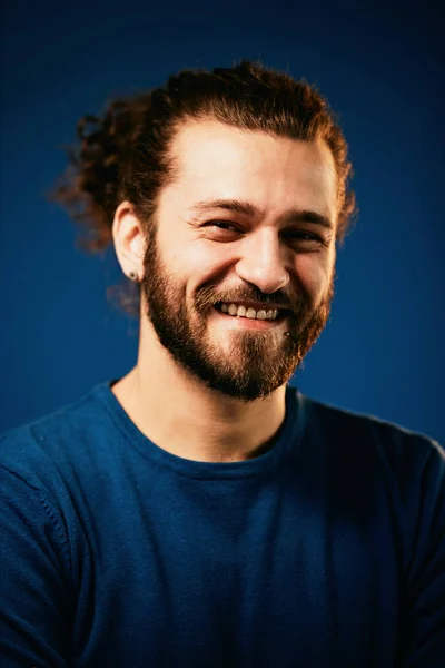 Young charming attractive Caucasian bearded hipster with curly hair dressed in sweater standing in front of blue background and looking at camera. — Stock Photo, Image