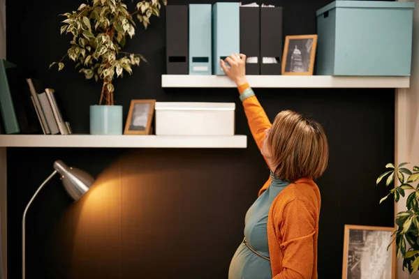 Preciosa mujer de negocios embarazada de pie en la oficina, tocando el vientre y tratando de llegar a aglutinante con documentos importantes . — Foto de Stock