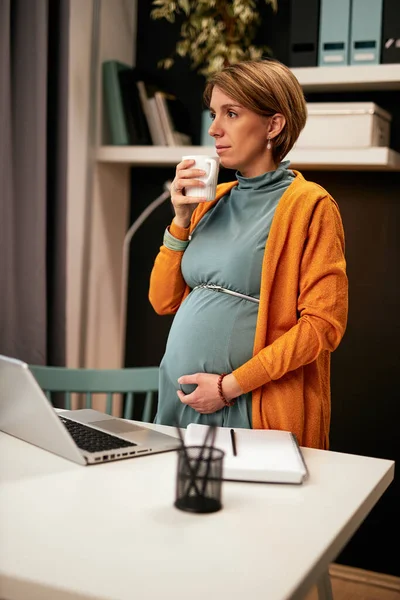 Charming relaxed caucasian pregnant accountant standing in her office, touching belly, drinking tea and looking trough window. — 스톡 사진