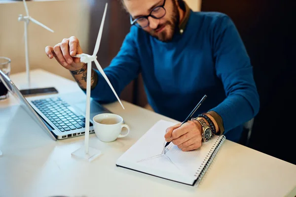 Junger kreativer kaukasischer bärtiger Umweltschützer mit lockigem Haar sitzt in seinem Büro, berührt Windmühlenmodell und zeichnet neues besseres Modell in Notizbuch. — Stockfoto