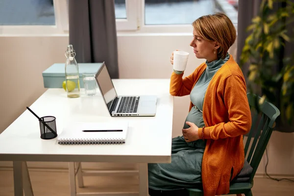Όμορφη καυκάσια έγκυος επιχειρηματίας στα σαράντα κάθεται στο γραφείο της, αγγίζοντας την κοιλιά και πίνοντας γλυκό νερό. — Φωτογραφία Αρχείου
