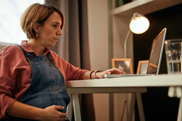 40 'lı yaşlarda beyaz, güzel, hamile bir kadın ev ofisinde oturuyor, karnına dokunuyor ve annelikle ilgili makaleler yazmak için dizüstü bilgisayar kullanıyor.. — Stok fotoğraf