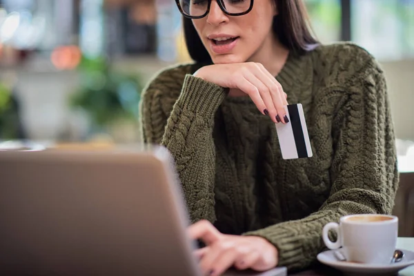 Attraktiv kaukasisk brunett i tröjan sittande på caféet, skriva på tangentbord och hålla kreditkort. — Stockfoto