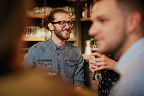 Bonito homem hipster sorridente sentado no pub perto do balcão do bar, segurando uma caneca de cerveja e ouvindo histórias interessantes. Vida noturna . — Fotografia de Stock