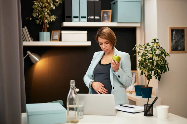 Attraktiv kaukasisk gravid affärskvinna i fyrtioårsåldern stående på kontoret, äta äpple och titta på laptop. — Stockfoto