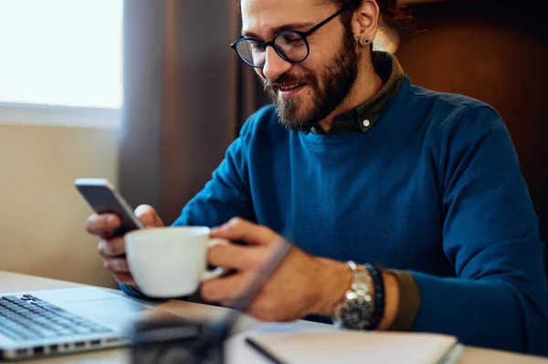 Joven hombre de negocios caucásico bien parecido sentado en su oficina, sosteniendo una taza de café y mirando el teléfono inteligente. Enfoque selectivo en el teléfono inteligente . — Foto de Stock