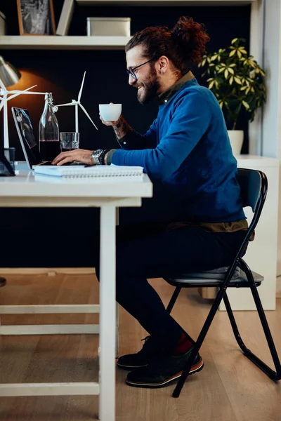 Charmig kaukasisk frilansare sitter i sitt hemmakontor, skriva på laptop rapport och hålla kopp kaffe. — Stockfoto