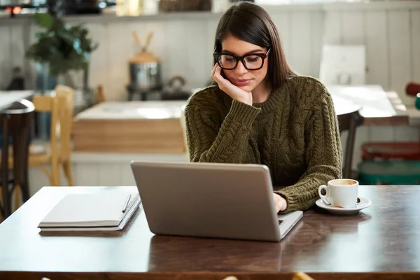 Charmante kaukasische Brünette im Pullover sitzt im Café und benutzt Laptop. — Stockfoto