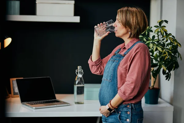 Πλευρική άποψη του χαριτωμένο έγκυος καυκάσιος γυναίκα στέκεται στο γραφείο στο σπίτι και πίνοντας γλυκό νερό. — Φωτογραφία Αρχείου