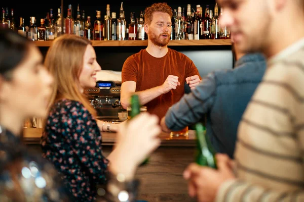 Vrolijke vrienden leunend op een bar, bier drinkend en babbelend met de barman. Avond uit.. — Stockfoto