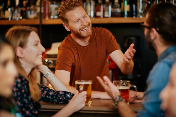 Veselí přátelé opírající se o bar, popíjející pivo a klábosící s barmanem. Noc.. — Stock fotografie