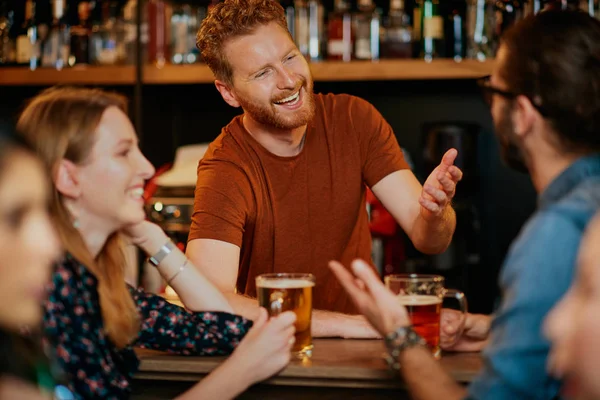 Vrolijke vrienden leunend op een bar, bier drinkend en babbelend met de barman. Avond uit.. — Stockfoto