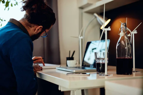 Joven e innovador empleado barbudo dedicado sentado en su moderna oficina y dibujando bocetos de molinos de viento. Concepto de desarrollo sostenible. —  Fotos de Stock