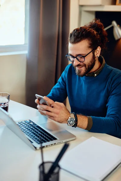 Glimlachende blanke hipster zit in zijn kantoor en gebruikt smartphone voor het beantwoorden van een e-mail. — Stockfoto