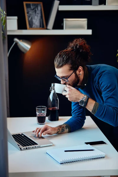 Jonge aantrekkelijke bebaarde hipster met krullend haar leunend op tafel, het drinken van koffie en surfen op internet tijdens het staan in huis kantoor. — Stockfoto