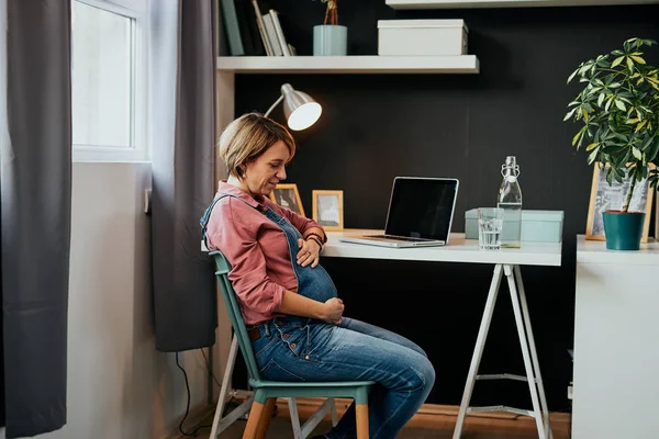 Stolta framtida mor sitter på sitt hemmakontor och vidrör magen. — Stockfoto