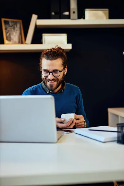 Jonge glimlachende knappe blanke freelancer zittend in zijn moderne kantoor thuis en met een kopje koffie. — Stockfoto