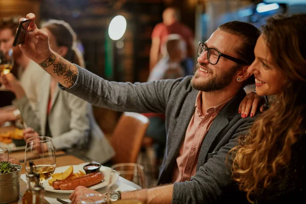 Joli couple souriant caucasien amoureux assis au restaurant et prenant selfie. Autour d'eux sont leurs amis bavarder et dîner . — Photo