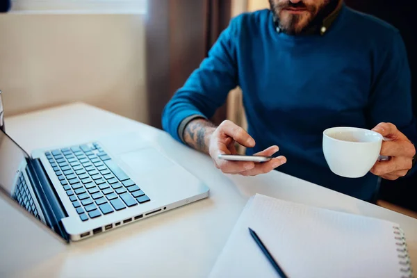 Giovane bel hipster barbuto seduto a casa sua ufficio, tenendo in mano una tazza di caffè e utilizzando lo smartphone. — Foto Stock