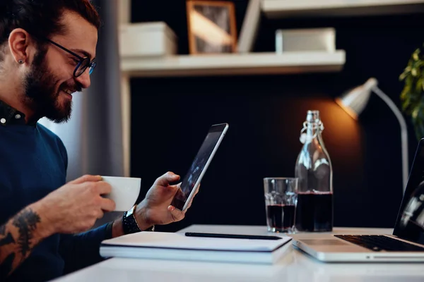 Νέοι ελκυστική καυκάσιος γενειοφόρος γραφίστας κάθεται στο γραφείο, πίνοντας καφέ και χρησιμοποιώντας tablet. — Φωτογραφία Αρχείου