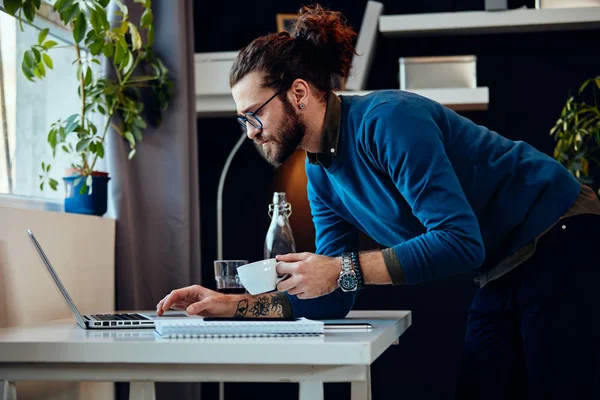 Ung attraktiv skäggig hipster med lockigt hår lutande på bordet, dricka kaffe och surfa på internet när du står på hemmakontoret. — Stockfoto