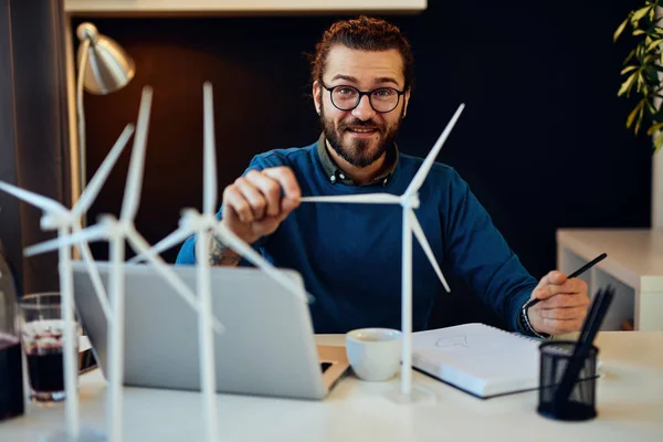 Young creative caucasian bearded environmentalist  with curly hair sitting in his office, touching windmill model and drawing new better model in notebook.