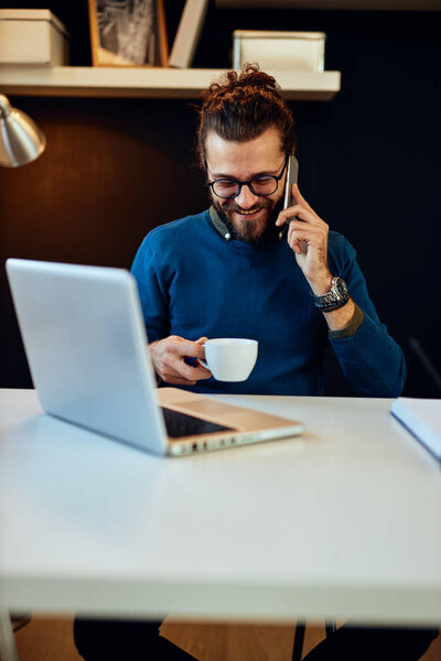 Young laughing caucasian bearded blogger sitting in his office, talking on the phone, holding cup of coffee and taking a break from work.