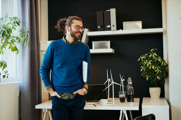 Knappe positieve jonge Kaukasische hipster staat in zijn kantoor, onder de oksel en voor te bereiden om te gaan op een ontmoeting met belangrijke klanten. — Stockfoto